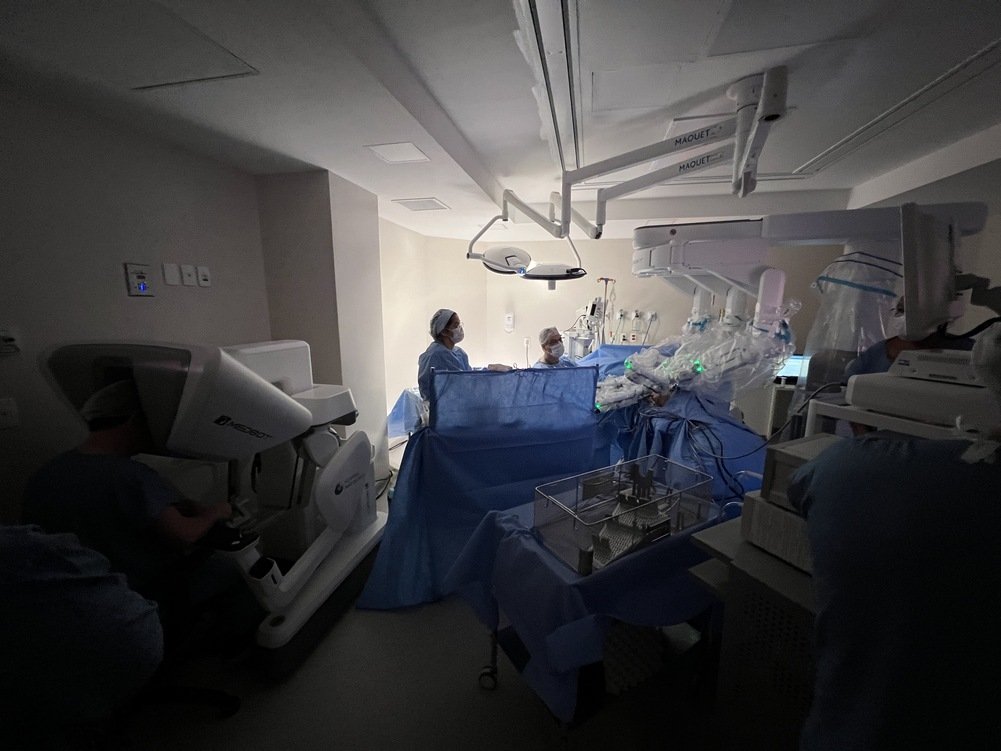 Sala cirúrgica com equipe médica durante procedimento robótico, com profissionais paramentados ao redor do paciente e braços do robô cirúrgico posicionados sobre a mesa, ilustrando artigo sobre cirurgia robótica Porto Alegre.