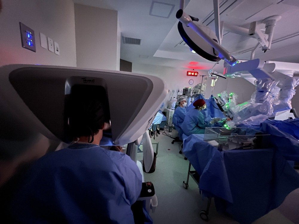 Profissional operando console robótico em primeiro plano enquanto equipe médica acompanha procedimento em sala cirúrgica com braços robóticos acoplados ao paciente, ilustrando artigo sobre cirurgia robótica Porto Alegre.