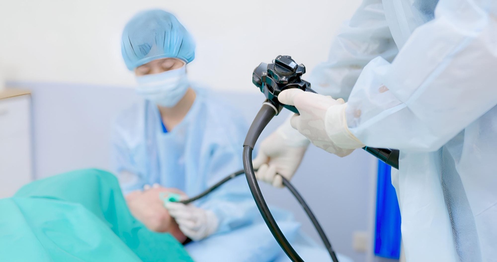 Profissional de saúde com luvas segura um equipamento de endoscopia ao lado de paciente deitado em sala de exame, com outro profissional ao fundo usando touca e máscara, ilustrando artigo sobre diferença entre endoscopia e colonoscopia