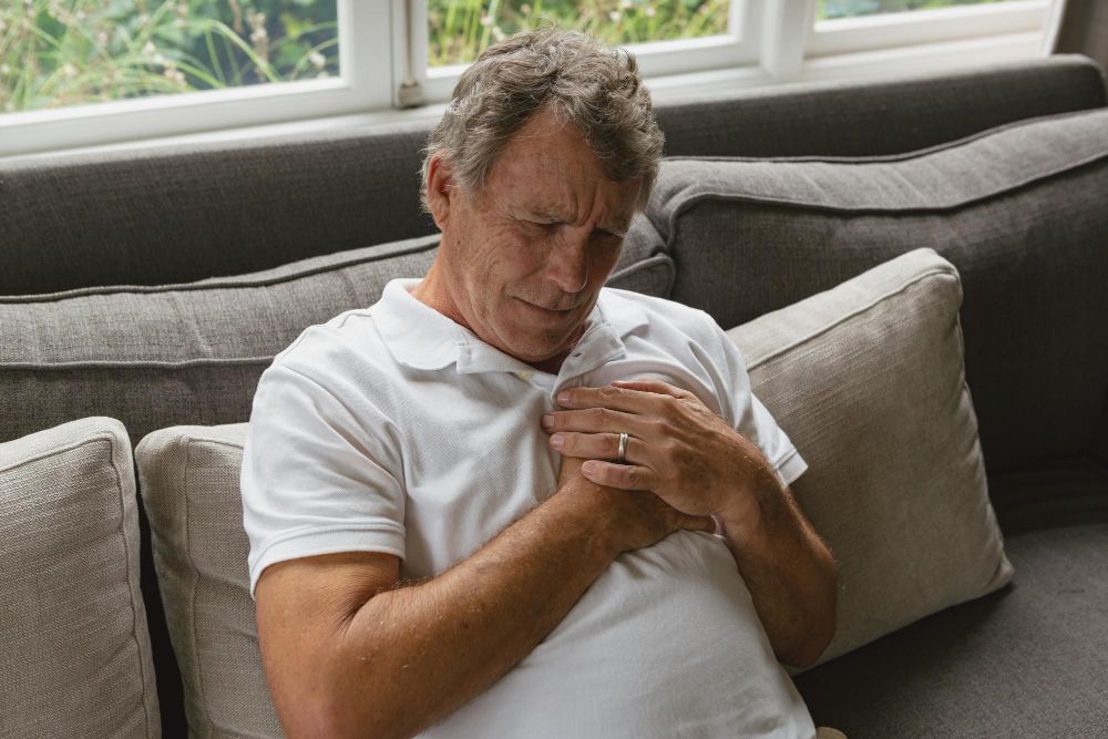 Médico de jaleco examinando o tórax de um paciente idoso sentado em maca de consultório, usando estetoscópio, ilustrando artigo sobre parada cardíaca o que fazer.