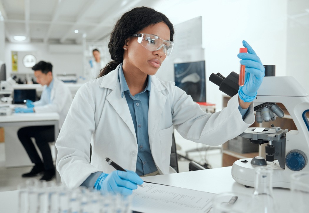 Profissional de laboratório com jaleco, luvas e óculos de proteção analisando tubo de ensaio ao lado de microscópio, ilustrando conteúdo sobre exames laboratoriais de rotina