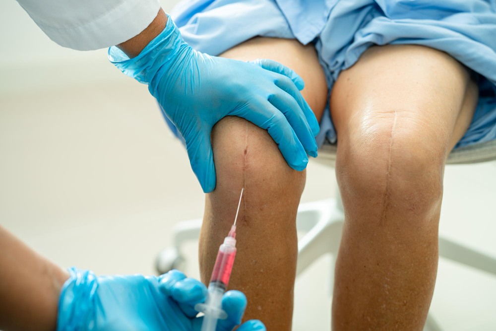 Profissional de saúde com luvas realizando infiltração no joelho de um paciente sentado, com agulha posicionada na articulação, ilustrando artigo sobre infiltração no joelho como funciona.