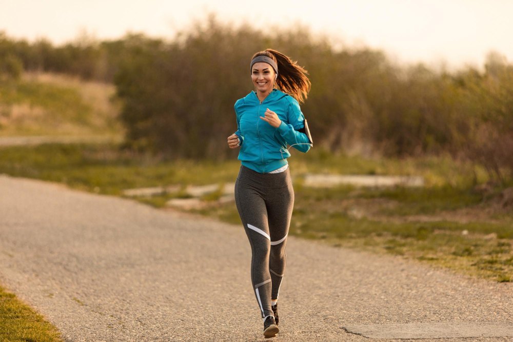 Mulher correndo em estrada ao ar livre usando roupas esportivas, representando prática de atividade física e risco de sobrecarga muscular, ilustrando artigo sobre lesões esportivas