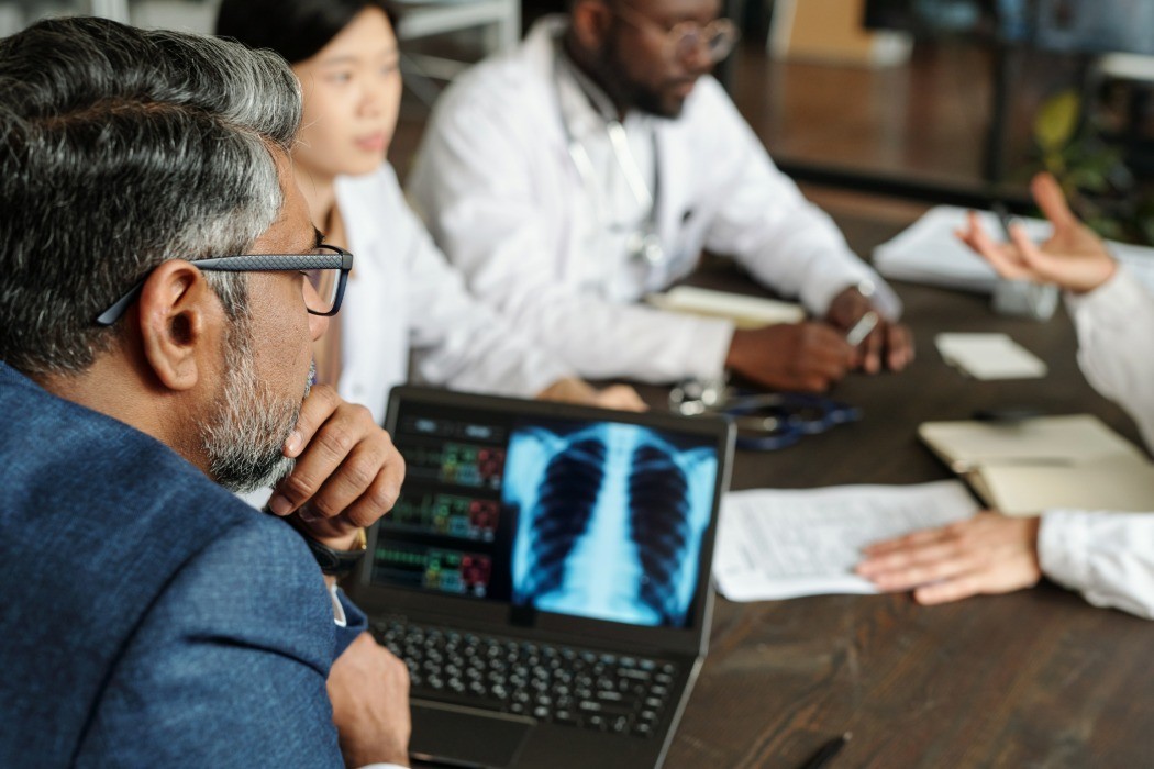 Médicos reunidos em torno de mesa discutindo caso, com radiografia de tórax exibida na tela de um laptop em primeiro plano, ilustrando conteúdo sobre nódulo pulmonar