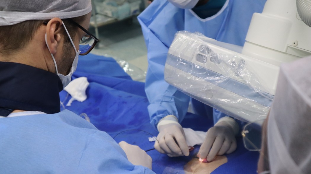 Equipe médica com aventais, luvas, máscaras e gorros realiza procedimento em paciente coberto por campos cirúrgicos azuis, com equipamento de imagem próximo ao local da punção, ilustrando artigo sobre o que é biopsia guiada por imagem.