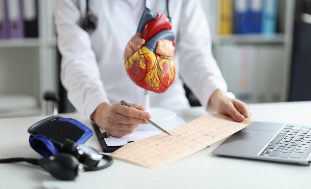 Médico de jaleco branco segura modelo anatômico do coração e aponta com caneta para exame de eletrocardiograma sobre a mesa, ao lado de aparelho de pressão arterial e notebook, ilustrando avaliação de palpitação no coração