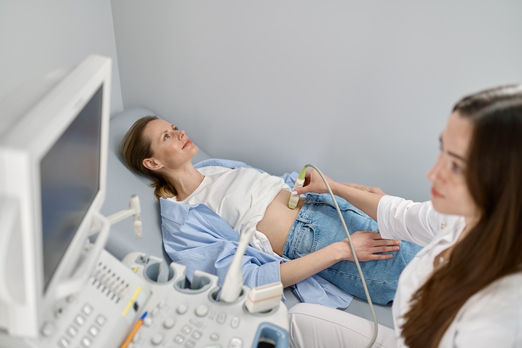 "Mulher deitada realizando ultrassom abdominal para diagnóstico de pedra na vesícula, com médica segurando o transdutor sobre o abdômen