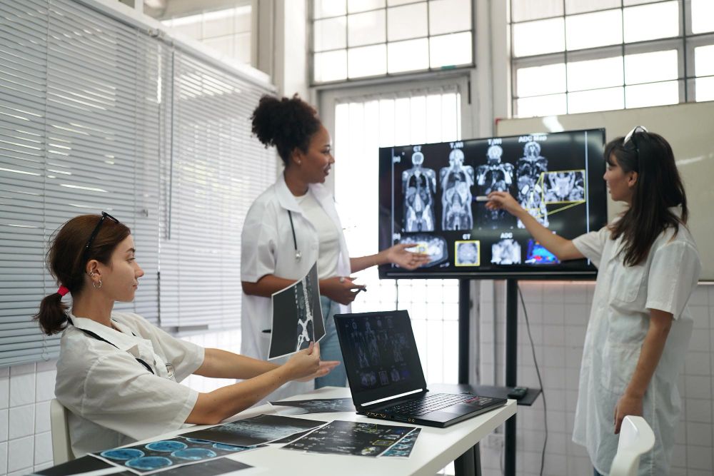 Três profissionais de saúde em uma sala de discussão clínica analisam exames de imagem em um monitor grande e em impressões sobre a mesa, ilustrando artigo sobre protocolos de exames em tomografia computadorizada e ressonância magnética.