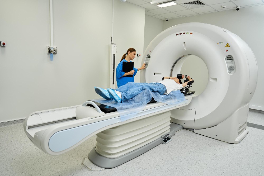 Paciente deitado em aparelho de tomografia computadorizada enquanto uma profissional de saúde acompanha o exame ao lado do equipamento em ambiente hospitalar, ilustrando artigo sobre protocolos de exames em tomografia computadorizada e ressonância magnética.