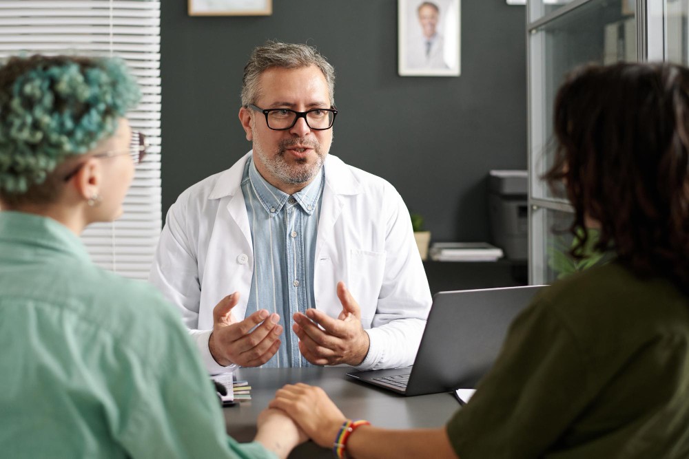 Médico conversa com duas pessoas durante consulta em consultório, com notebook sobre a mesa e ambiente clínico ao fundo, ilustrando artigo sobre quando procurar um oncologista 