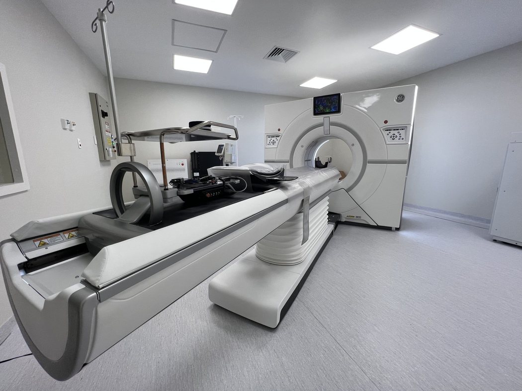 Sala de exames moderna e iluminada com um aparelho de tomografia de tórax de última geração. O tomógrafo possui formato circular branco e está conectado a uma maca hospitalar motorizada, preparada para acomodar o paciente de forma confortável em um ambiente médico limpo e humanizado.
